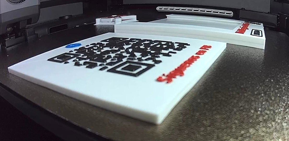 Un soporte y una placa con código QR de ScaleMint Tap en plena impresión 3D, con el QR en negro, el logo en azul y las letras en rojo